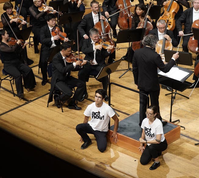 Die beiden Aktivisten kleben sich am Dirigentenpodest fest, während das Orchester weiterspielt.